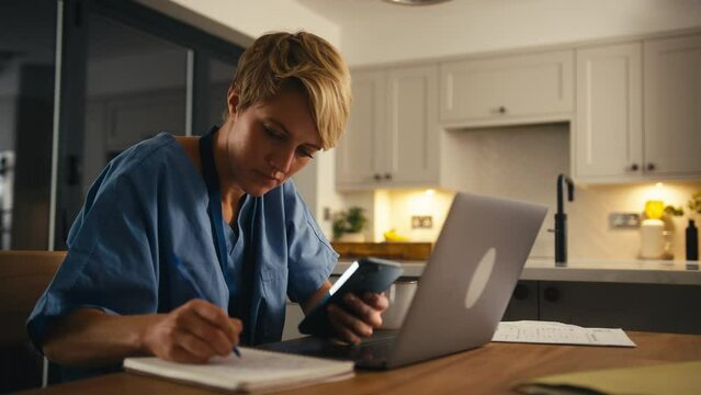 Tired woman wearing medical scrubs looking at mobile phone whilst working or studying on laptop at home at night - shot in slow motion