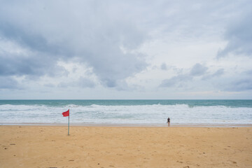 Female tourist standing on the beach, strong white waves and blue sky with white clouds, red flag symbolizing prohibition of swimming in the sea.