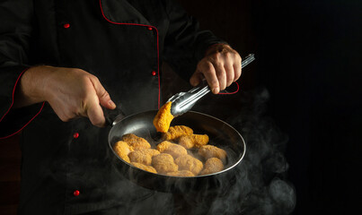 Chef prepares chicken nuggets in a frying pan. The concept of cooking nagits in the kitchen of a restaurant or hotel. Free space for advertising on a black background