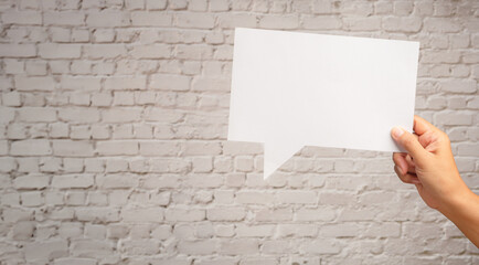 Hand holding a blank white speech bubble while standing against a white brick wall background.