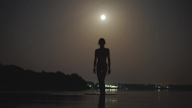 silhouette of slim woman in bikini walking in shallow water in lake at night with moonlight. summertime night sensual girl swimming in darkness