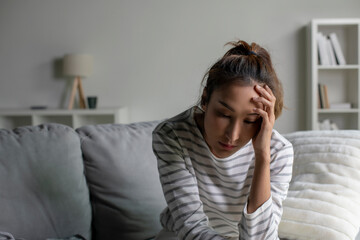 Lonely Asian woman in distress, worried, confused, sitting on sofa at home. Female suffer from mental health problems.