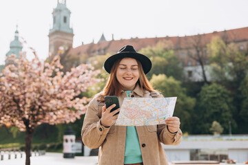 Attractive young female tourist is exploring new city. Redhead 30s girl holding a paper map and smart phone in Krakow. Traveling Europe in spring. High quality photo