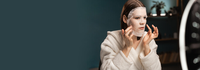 Young female beautician making beauty tutorial on smartphone at home. Female blogger is applying facial sheet mask on face. Cosmetic procedures concept.