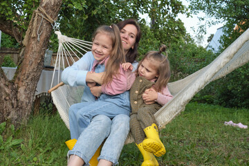 A joyful mother hugs her cheerful little daughters while relaxing in a hammock, showcasing a tranquil backyard momen