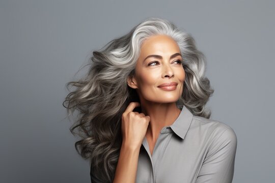 Adult Woman Touches Face With Smooth Healthy Skin. Beautiful Aging Mature Woman With Long Gray Hair And Happy, Shy Smiles On A Gray Background.