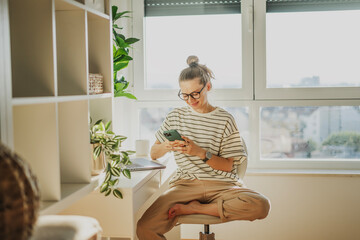 Young adult Caucasian woman in glasses working on a laptop and talking using a smartphone in a modern apartment office near the window