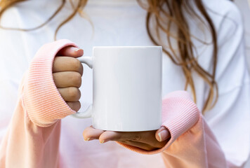  Girl is holding blank 11 oz white mug in hands and pink- white sweatshirt . Blank ceramic cup in hands