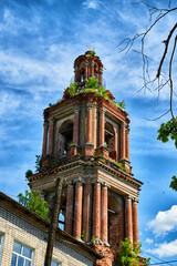 Fototapeta na wymiar A tall old stone bell tower made of red brick against a blue sky with white clouds and green trees around. Partially destroyed ancient bell tower and tower on a summer day