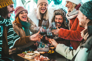 Happy friends group toasting red wine dining at restaurant terrace - Young people socializing drinking and eating food sitting outside at winery bar table - Winter season - Dinner lifestyle concept