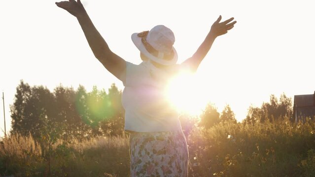 Elderly woman in hat and dress is turning around spot with pleasure raising arms. Outdoors on summer evening against rays of sun at sunset. Joy of freedom and positive emotions