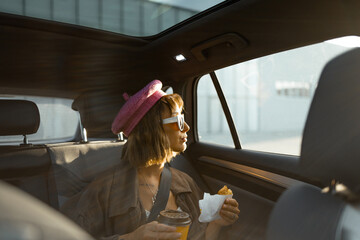 Young stylish woman sitting with a coffee cup and croissant on backseat of modern taxi while driving at city at sunny evening. Fast food and transportation concept