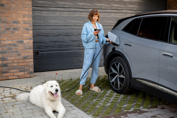 Young woman stands with her dog and using smart phone while charging her electric car near garage...