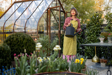 Portrait of a young woman as florist taking care of flowers and plants in garden with vintage greenhouse on background. Concept of hobby and gardening