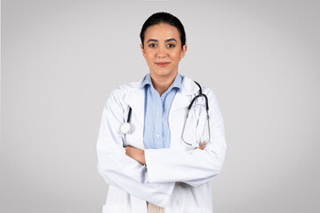 Fototapeta premium Smiling latin doctor in white uniform posing with arms folded in studio
