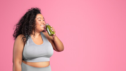 Black chubby woman in sportswear drinking bottle of juice or smoothie, pink studio background,...