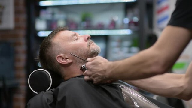 Barber master shaving handsome mature bearded man using electric shaver in salon. Hair artist making beard style for person in male barbershop. Services of professional stylist. Fashion haircare for m