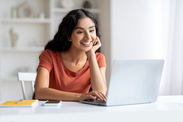 Pretty young indian woman gently smiles while browsing on laptop