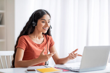 Cheerful remote worker pretty indian woman using laptop and headset
