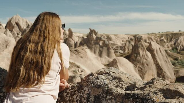 The young woman with long hair, leaning against a rock, admires the ancient stones from a height in the Valley of Imagination.