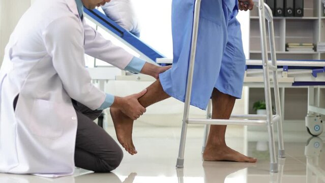 Doctor in a hospital is helping a male patient with weak leg muscles learn to walk with a cane. Physiotherapist doing physiotherapy for man practicing walking in the hospital room.