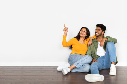 Happy young indian couple buying new house, sitting on floor in empty room, pointing at free space over white wall