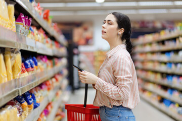 Side view of portrait of pretty caucasian woman holds smartphone and shopping cart. Choice products in supermarket