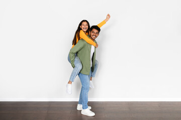 Overjoyed young indian man giving piggyback ride to his wife, posing over white wall background,...