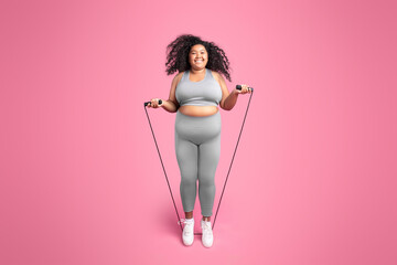 Happy african american chubby woman in sportswear jumping on jump rope, isolated on pink studio background, full length - obrazy, fototapety, plakaty