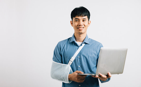 Confident Asian businessman with a broken arm, wearing a splint after an accident, continues working, holding a laptop. Studio isolated on white with copy space, highlighting resilience and recovery.