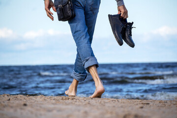 On the seashore of blue color, a man in denim pants walks on the beach sand barefoot and with black...