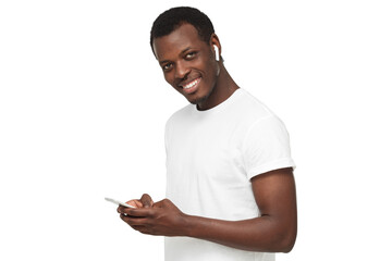 Young african american man looking and smiling at the camera, using mobile phone, listening to music with white wireless earphones