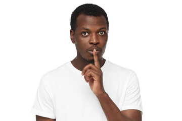 Fototapeta na wymiar Shh! Young african american man wearing blank white t-shirt and pressing index finger to lips as if asking other to keep silent about his secret