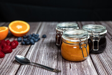 Assortment of homemade jams. Jams in glass jars, oranges, strawberries and blueberries. Front view with black background, seasonal fruit jam, marmalade, teaspoon.