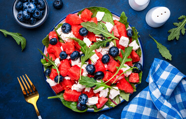 Fresh watermelon salad with soft goat cheese, blueberries and arugula in plate, blue table background, top view