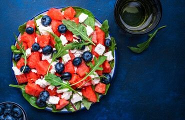 Fresh watermelon salad with soft goat cheese, blueberries and arugula in plate, blue table background, top view