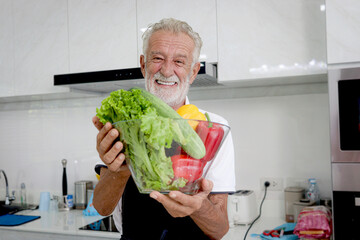 Elderly man standing at counter holding colorful fresh vegetables bowl for making salad, senior man cooking healthy food with organic ingredients at home, mature grandfather making meal at kitchen.