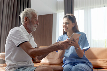 Female nurse takes care of senior patient and teaches him to use digital tablet at living room house, elderly mature grandfather learns to online internet and high-tech, nursing medical health care.