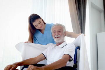 Obraz premium Beautiful female nurse takes care of senior patient in wheelchair at home, disabled elderly man sits in wheelchair while woman doctor covers blanket to mature grandfather, nursing medical health care.