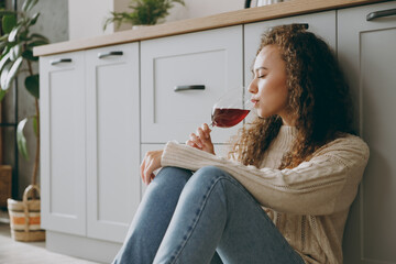 Close up young calm happy fun cool housewife woman of African American ethnicity wear casual clothes sweater drink wine sit near table in light kitchen at home alone. Lifestyle cooking food concept. - obrazy, fototapety, plakaty