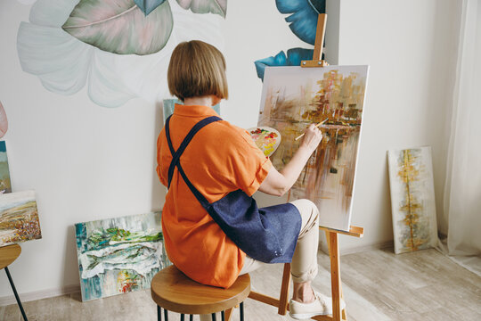Full body back rear view elderly artist woman 50 years old wearing casual clothes sitting near easel with painting artwork paint spending free spare time in living room indoor. Leisure hobby concept.