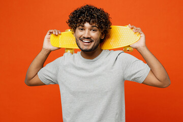 Young smiling cheerful cool happy Indian man he wears t-shirt casual clothes hold yellow pennyboard skateboard loking camera isolated on orange red color background studio portrait. Lifestyle concept. - obrazy, fototapety, plakaty