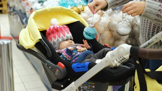 Mother showing Xmas balls to her baby at a store. Cute infant girl lying in the stroller while shooping for Christmas decorations