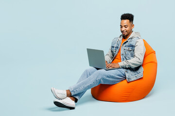 Full body young IT man of African American ethnicity wear denim jacket orange t-shirt sit in bag chair hold use work on laptop pc computer isolated on plain pastel light blue cyan background studio.