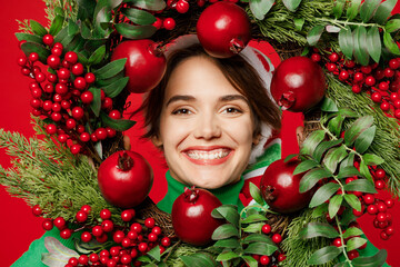 Close up young fun woman wear green turtleneck Santa hat posing hold in hand look through decorative wreath isolated on plain red background. Happy New Year 2024 celebration Christmas holiday concept.