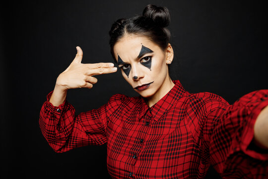Young creepy woman with Halloween makeup face art mask wear clown costume red dress doing selfie shot pov mobile cell phone isolated on plain solid black background studio Scary holiday party concept.