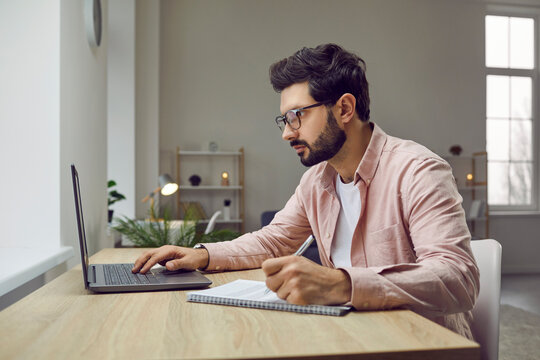 Side view shot of young man wearing glasses using laptop. Businessman sitting at desk looking at computer screen and writing down list to do in notepad in comfortable office