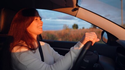 Cheerful young woman drives modern car along rural road at sunset light
