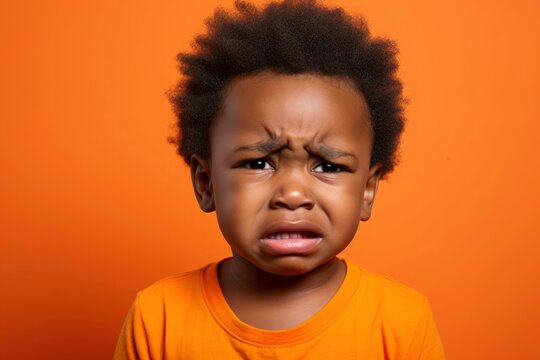 Dark-skinned African-American Todler child with curly hair in an orange T-shirt is crying on an orange background. Upset kid, sad, frowned, angry. Concept of people's emotions, child crisis