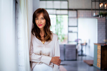 Portrait of a brunette haired middle-aged woman wearing a shirt and pants and standing by the window